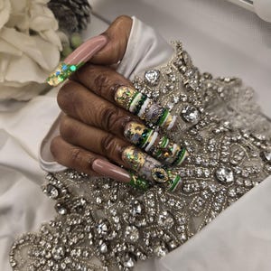Op de afbeelding: Lange vierkante kunstnagels met een complex ontwerp met een nude basis, groene accenten, bladgoud, witte strepen en ingebedde strass-steentjes. De nagels zijn versierd met ingewikkelde patronen, metallic gouden details en kleine groene edelsteentjes.