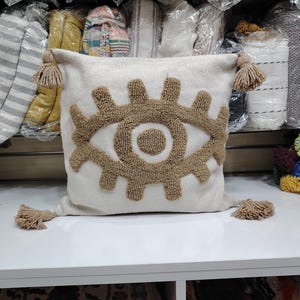 Peut inclure: Coussin décoratif crème avec un grand motif d'œil marron brodé. Le coussin a des glands assortis à chaque coin. Le coussin est posé sur une surface blanche.