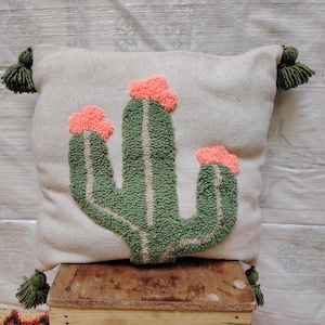 Peut inclure: Un oreiller blanc avec un motif de cactus vert. Le cactus a trois bras et des fleurs roses. L'oreiller a des glands verts aux coins.