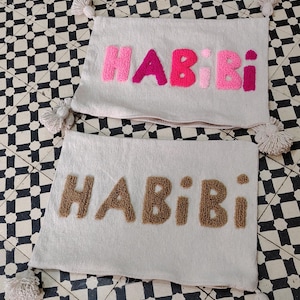 Peut inclure: Deux coussins décoratifs blanc cassé avec le mot "HABIBI" en lettres texturées roses et marron. Chaque coussin a un pompon à chaque coin. Les coussins sont sur une surface à carreaux noirs et blancs.