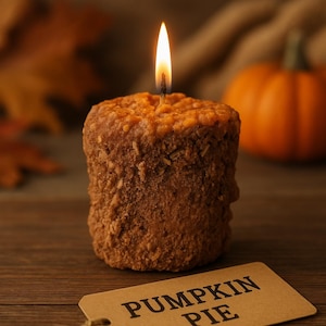 May include: A lit pumpkin pie-shaped candle with a textured, brown exterior and a textured, orange top. A small orange pumpkin is in the background. A tag that says "PUMPKIN PIE" is in the foreground.