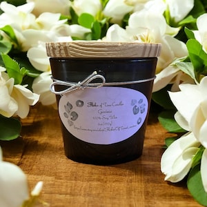 Könnte beinhalten: Ein schwarzes Kerzenglas mit Holzdeckel und silberner Schleife. Das Etikett lautet "Picker of Time Candles, Gardenia, 100% Soy Wax, 170g". Die Kerze ist von weißen Blumen umgeben.