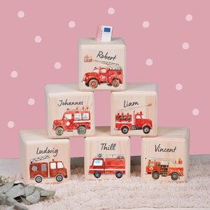 Könnte beinhalten: Holzblöcke mit roten Feuerwehrwagen-Illustrationen und Namen: Robert, Johannes, Liam, Ludwig, Thill und Vincent. Ein Geldschein steckt im obersten Block. Der Hintergrund ist rosa mit weißen Punkten.