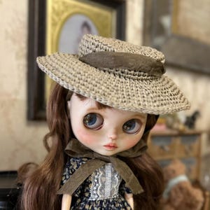 以下が含まれることがあります： 大きな青い目と長い茶色の髪の毛を持つ人形。茶色のバンドが付いた麦わら帽子をかぶっています。人形は、首に茶色のリボンが付いた濃い青色の花柄のドレスを着ています。背景には額入りの写真とぼやけたインテリアが含まれています。