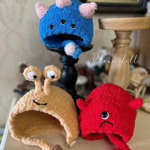 以下が含まれることがあります： 青、ベージュ、赤の3色の手作りのモンスターハット。青い帽子には3つの目とピンクの角があります。ベージュの帽子には2つの大きな目と笑顔の口があります。赤い帽子には1つの目と悲しい口があります。