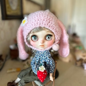 以下が含まれることがあります： 白い花があしらわれたピンクの編みぐるみウサギの帽子。青い目をした人形が花柄のドレスを着て帽子をかぶっています。人形は赤と白のイチゴを持っています。