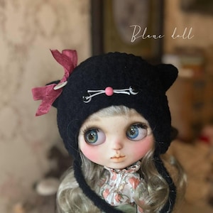 以下が含まれることがあります： 黒いかぎ針編みの猫耳帽子をかぶった人形。ピンクの鼻、白いひげ、サイドにダークレッドのリボンが付いています。人形は大きな青い目、色白の肌、長いウェーブのかかった髪をしています。右上に「Blanc doll」という文字が見えます。