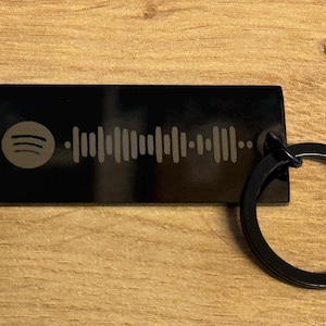 Könnte beinhalten: Ein schwarzer, rechteckiger Schlüsselanhänger mit einem Spotify-Code und -Logo. Der Schlüsselanhänger hat einen schwarzen Schlüsselring. Das Spotify-Logo befindet sich oben links, der Code in der Mitte. Der Schlüsselanhänger liegt auf einer Holzoberfläche.