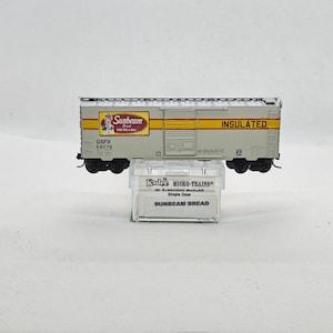 May include: A model train boxcar with the Sunbeam Bread logo and the word "INSULATED" printed on the side. The boxcar is gray with yellow stripes and black wheels. It sits on a clear plastic display case.