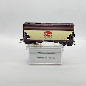 May include: A model train car with a brown roof and sides, and a cream-colored center panel. The panel features the "Frostie Root Beer" logo and text. The car is on a clear plastic display case.