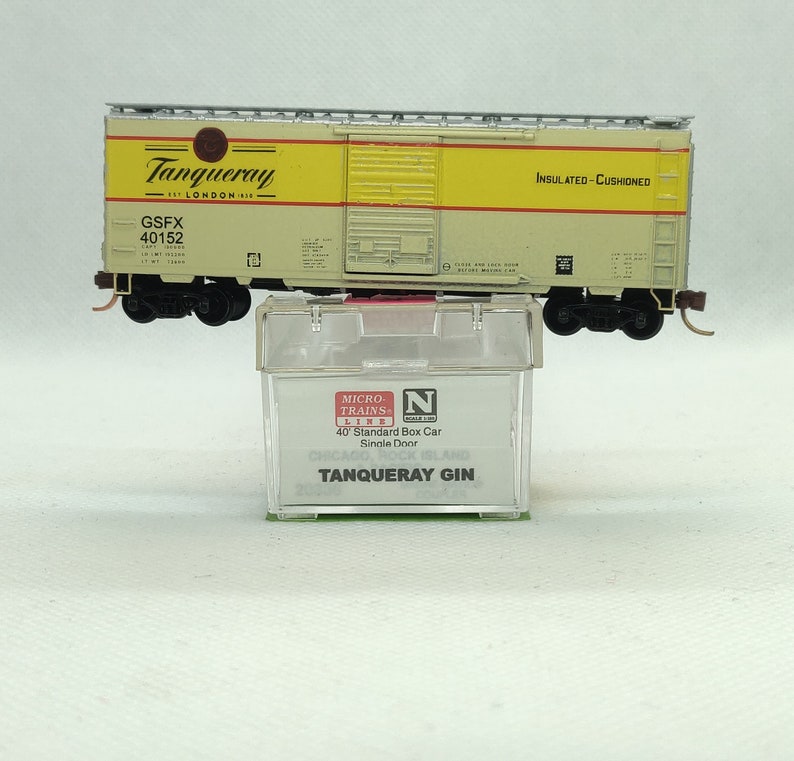 Pode incluir: Um vag&atilde;o de mercadorias modelo com as palavras "Tanqueray London Gin" e "Insulated Cushioned" impressas no lado. O vag&atilde;o est&aacute; numa vitrine de pl&aacute;stico transparente com as palavras "Micro-Trains Line 40' Standard Box Car Single Door" e "Tanqueray Gin" impressas na vitrine.