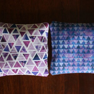 May include: Two square pillows with geometric triangle patterns. One pillow features a white background with purple and blue triangles. The other pillow has a blue background with purple triangles.