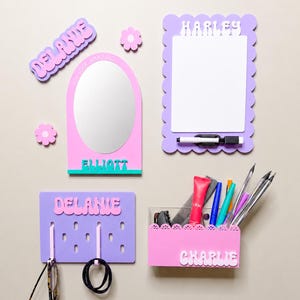 May include: A collection of personalized wall-mounted items including a mirror with the name ELLIOTT, a scalloped whiteboard labeled HARLEY with a marker, a purple pegboard labeled DELANIE, and a pink storage bin labeled CHARLIE holding various items.