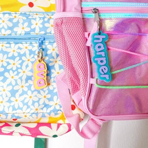 May include: Close-up of two colorful backpacks. One backpack has a yellow background with white flowers and a blue zipper. The other is pink with a holographic sheen and a teal zipper. Each has a name tag: "ava" and "harper".