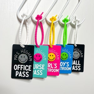 May include: Five colorful plastic name tags with a smiley face design and the text "Mrs. Higham Office Pass", "Mrs. Higham Nurse Pass", "Mrs. Higham Girl's Room", "Mrs. Higham Boy's Room", and "Mrs. Higham Hall Pass".