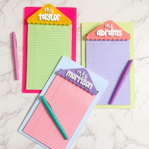 Op de afbeelding: Drie gepersonaliseerde notitieblokken met pennen. Elk notitieblok heeft papier in een andere kleur en een decoratieve bovenkant met een naam. De kleuren van de notitieblokken zijn roze, groen en paars. De pennen zijn roze, paars en blauwgroen.