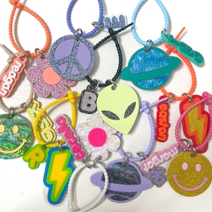 May include: A collection of colorful acrylic charms with various designs, including a peace sign, smiley faces, flowers, planets, and lightning bolts. Each charm is attached to a matching colored loop. Some charms have names on them.