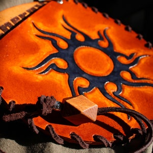 May include: A round, orange leather pouch with a black sun design and a brown leather strap. The pouch has a wooden bead on the strap.