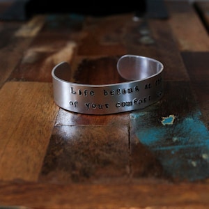 May include: A silver cuff bracelet with the stamped inscription "Life begins at the of your comfort zone." The open-ended bracelet is displayed on a wooden surface with a mix of brown, blue, and green tones.