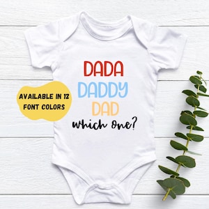May include: White baby bodysuit with the words "DADA," "DADDY," and "DAD" in red, blue, and yellow, respectively, with "which one?" in black. A yellow circle with the text "AVAILABLE IN 12 FONT COLOURS" is on the left.