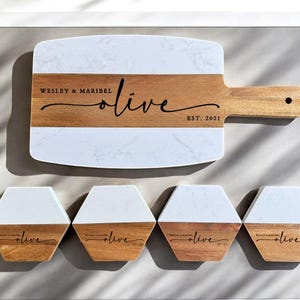 May include: A white and brown rectangular serving board with a marble design and a wooden handle. The board has the text "Wesley & Maribel olive EST. 2021" engraved on it. Four smaller white and brown hexagonal coasters with the same design and text are also shown.