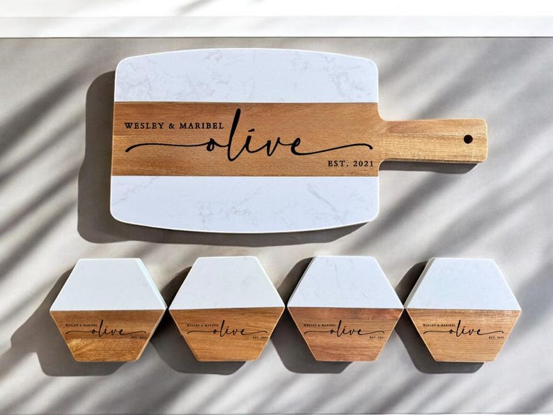 May include: A white and brown rectangular serving board with a marble design and a wooden handle. The board has the text "Wesley & Maribel olive EST. 2021" engraved on it. Four smaller white and brown hexagonal coasters with the same design and text are also shown.