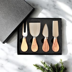 May include: A set of four cheese knives with wooden handles in a black box. The knives have different blade shapes, including a fork, a spreader, and two different types of knives.