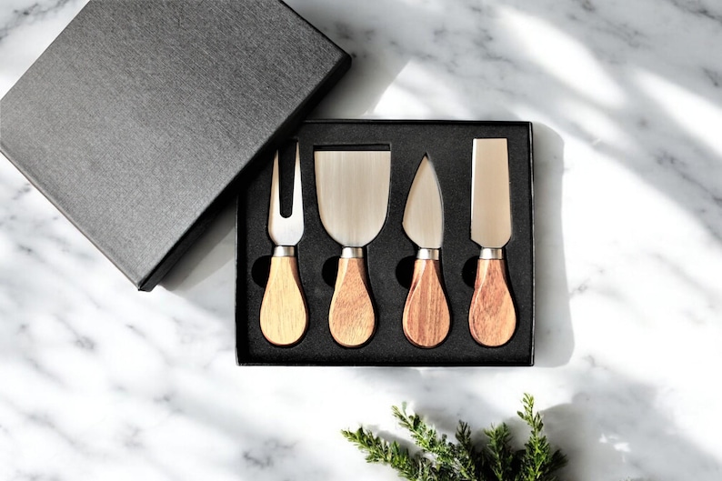 May include: A set of four cheese knives with wooden handles in a black box. The knives have different blade shapes, including a fork, a spreader, and two different types of knives.