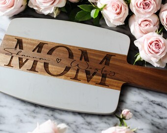 May include: A white and wood cutting board with the word "MOM" engraved in large letters. The words "Shannon" and "Wesley" are engraved in smaller letters below the word "MOM".