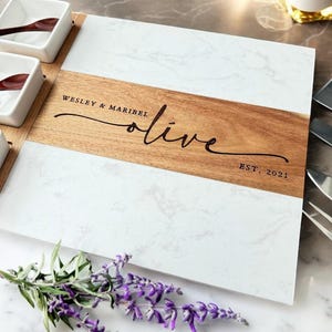 May include: A white marble and wood serving board with a personalized inscription: "Wesley & Maribel olive est. 2021". The board has three small white bowls and four serving utensils.