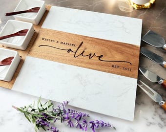 Puede incluir: Una tabla de servir de mármol blanco y madera con una inscripción personalizada: "Wesley & Maribel olive est. 2021". La tabla tiene tres cuencos pequeños blancos y cuatro utensilios de servir.