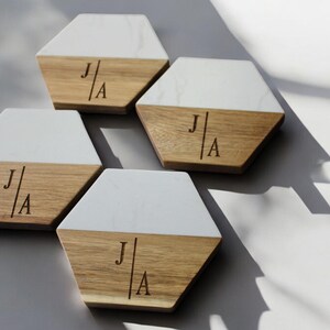 May include: Set of four hexagonal coasters made of wood and white marble. Each coaster has a white marble top and a wood bottom with the initials "J|A" engraved on the wood.