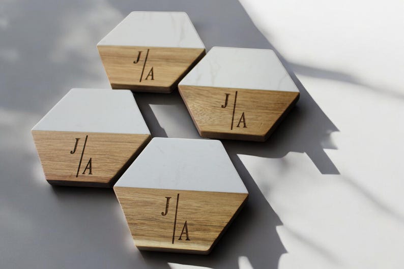 May include: Set of four hexagonal coasters made of wood and white marble. Each coaster has a white marble top and a wood bottom with the initials "J|A" engraved on the wood.