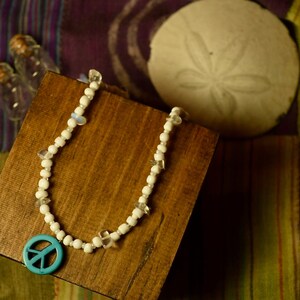 Puede incluir: Un collar de cuentas blancas con un dije de signo de paz turquesa. El collar se muestra en una caja de madera con un dólar de arena en el fondo.