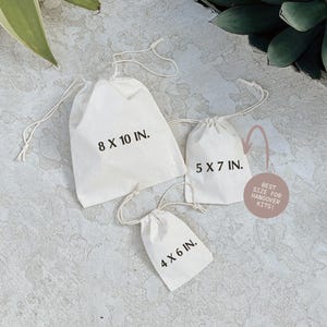 three white tote bags with measurements printed on them, placed on a textured surface.