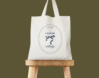 Puede incluir: Bolso tote color crema con un logotipo azul marino que presenta un lazo y las palabras "custom college" dentro de un óvalo. La bolsa está sobre un pequeño taburete de madera sobre un fondo verde apagado.