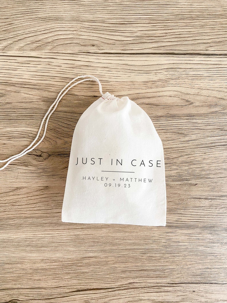 May include: A white cotton drawstring bag with the text "JUST IN CASE" printed in black. Below the text is "HAYLEY + MATTHEW 09.19.23" printed in black.