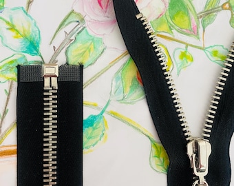 May include: Close-up of two black zippers with silver teeth and pulls. The zippers are set against a backdrop of a watercolour painting of pink and yellow roses with green leaves. One zipper is closed, and the other is open.