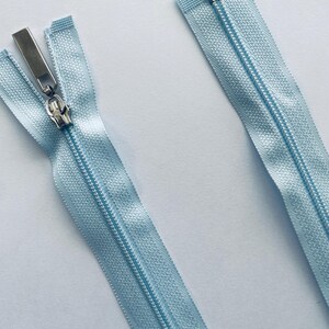 May include: Two pale blue zippers with silver metal pulls. One is closed, the other open, showing the teeth. The zippers are set against a white background.