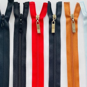 May include: A selection of colourful zips in various shades, including navy, white, black, red, brown, orange, light blue, and dark brown. Each zip is closed and features a metal pull tab. The zips are arranged in a row.