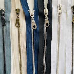 May include: Assortment of six zipper tapes with different colors and metal slider pulls. The colors include blue, black, white, and beige.