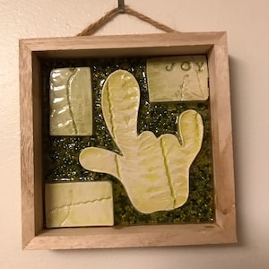 May include: A framed ceramic wall art piece with a green and white color scheme. The piece features a hand-shaped design with the word "JOY" in the upper right corner.