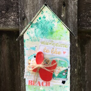 May include: A wooden house-shaped sign with a green, white, and blue watercolor background. The sign says "Welcome to the Beach" with a red seashell and a green and white van graphic.