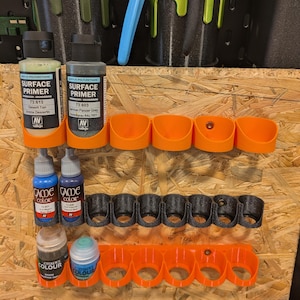May include: A collection of paint bottles and holders in various colors. The image shows bottles of primer and paint, along with orange and black holders. The holders are designed to organize the paint bottles. The background includes black containers holding pens and brushes.