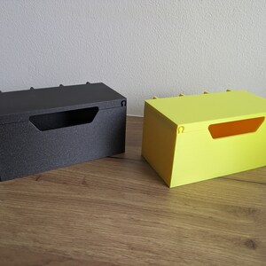 May include: Two small storage boxes, one black and one yellow, with a rectangular opening on the front. Both boxes have a textured surface and are made of plastic.