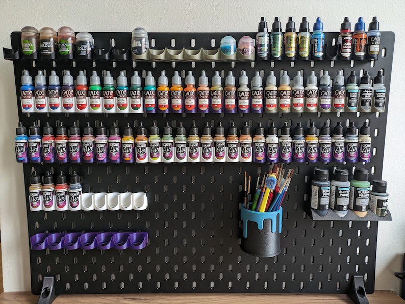 May include: A black pegboard with a variety of paint bottles organised in rows. The bottles are mostly small and have different coloured labels. There are also some paint brushes and a small cup for holding them.