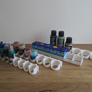 May include: White plastic paint bottle holder with 12 slots. The holder is filled with various bottles of paint, including black, blue, green, and red. The bottles are labelled with the words "Aqua Colour" and "Surface Primer".