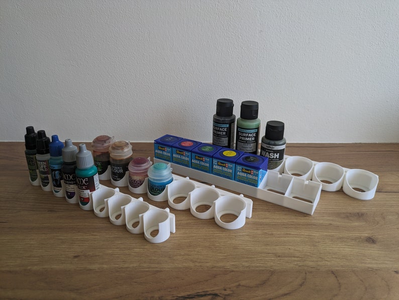 May include: White plastic paint bottle holder with 12 slots. The holder is filled with various bottles of paint, including black, blue, green, and red. The bottles are labelled with the words "Aqua Colour" and "Surface Primer".
