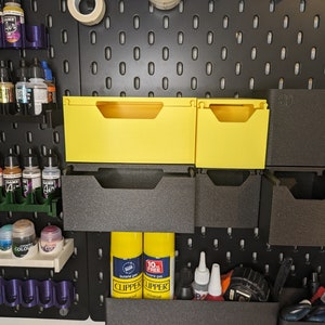 May include: A black pegboard wall with yellow and black storage bins. The bins are stacked on top of each other and are labeled with numbers. There are also various bottles of paint and other supplies on the wall.