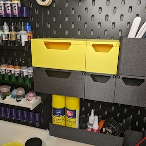 May include: A pegboard wall with various storage solutions, including yellow plastic drawers, black plastic bins, and a shelf with small bottles. The pegboard is black with a grid pattern.
